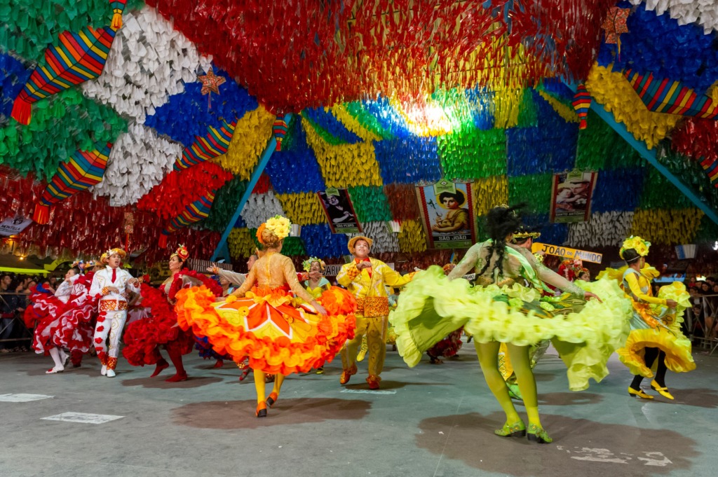 Apresentação de dança em festa junina dentro de um espaço coberto, com o teto totalmente decorado por bandeirinhas e tiras coloridas em vermelho, amarelo, azul, verde e branco. Vários dançarinos usam trajes típicos vibrantes, com saias rodadas em camadas de cores intensas como vermelho, amarelo e verde, girando durante a coreografia. Ao fundo, há cartazes com imagens e textos relacionados a São João, enquanto o público observa nas laterais do espaço.