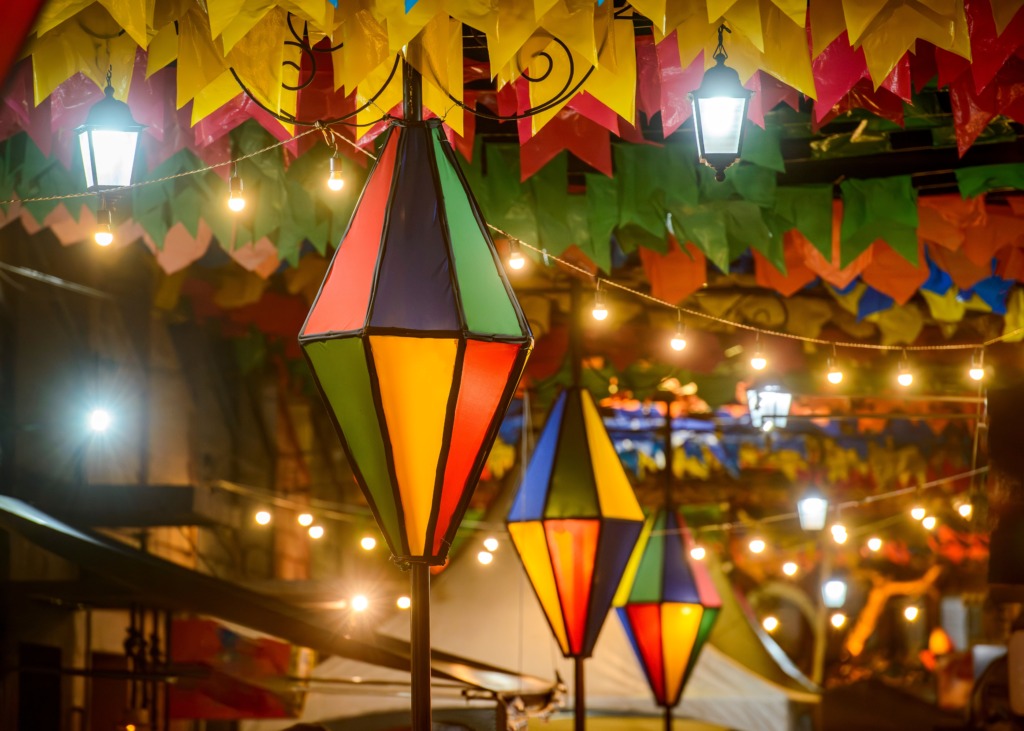 Detalhe de decoração de festa junina com lanternas coloridas penduradas em um ambiente noturno, cercadas por bandeirinhas de papel em amarelo, verde, vermelho e azul e luzes pequenas que iluminam o espaço, criando uma atmosfera festiva e acolhedora.