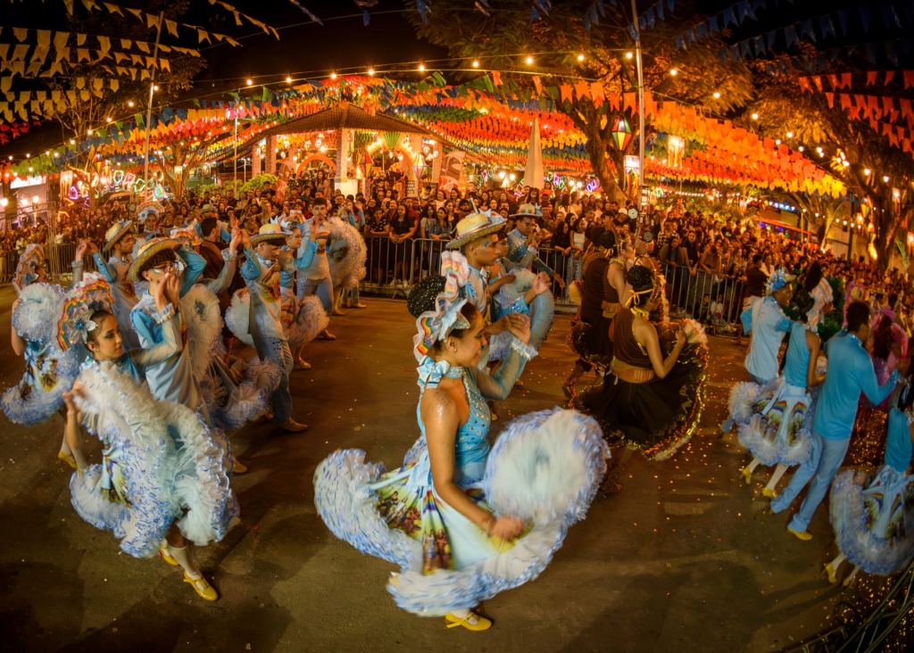 Festa junina noturna rua decorada com bandeirinhas coloridas e luzes, com várias pessoas dançando em trajes tradicionais com saias rodadas e chapéus, enquanto uma grande multidão observa atrás de grades.